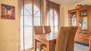 Un comedor con mesa de madera y sillas tejidas, ventanas amplias con persianas y un armario con vajilla.