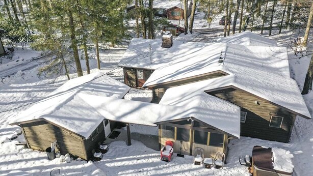 Una cabaña cubierta de nieve con techo inclinado, un cobertizo pequeño adosado y un porche cubierto con mobiliario exterior.