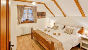 A bedroom with a wooden ceiling, a large bed with multiple pillows, a nightstand with a lamp, a window with curtains, and a radiator.