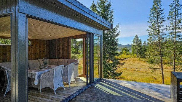 Una moderna cabina con una porta in vetro che si apre su un terrazza in legno, arredata con un divano e poltrone, con vista su un paesaggio boschivo.