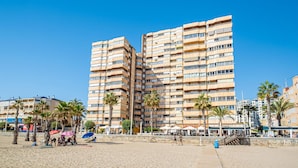 Un hotel frente a la playa con varios balcones, palmeras y servicios junto a la costa.