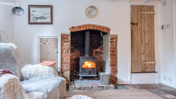 A cozy room with a brick fireplace, a wooden door, and a wicker chair. - Lilac Cottage, Wenhaston (Halesworth)