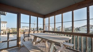 A wooden deck with a picnic table and benches, large windows offering a view of houses and a clear sky. - 7692- Seaside Serenity - Pet-Friendly Avon Retreat | Sunsets & Sun Decks (Avon)