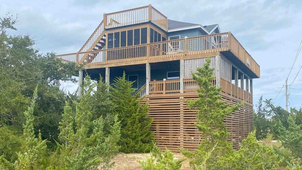 A multi-level wooden house with a balcony, surrounded by dense greenery. - 7692- Seaside Serenity - Pet-Friendly Avon Retreat | Sunsets & Sun Decks (Avon)