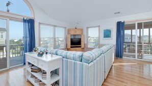 A spacious living room with a large window, a built-in TV, a striped sofa, and a white coffee table with books. - 7692- Seaside Serenity - Pet-Friendly Avon Retreat | Sunsets & Sun Decks (Avon)