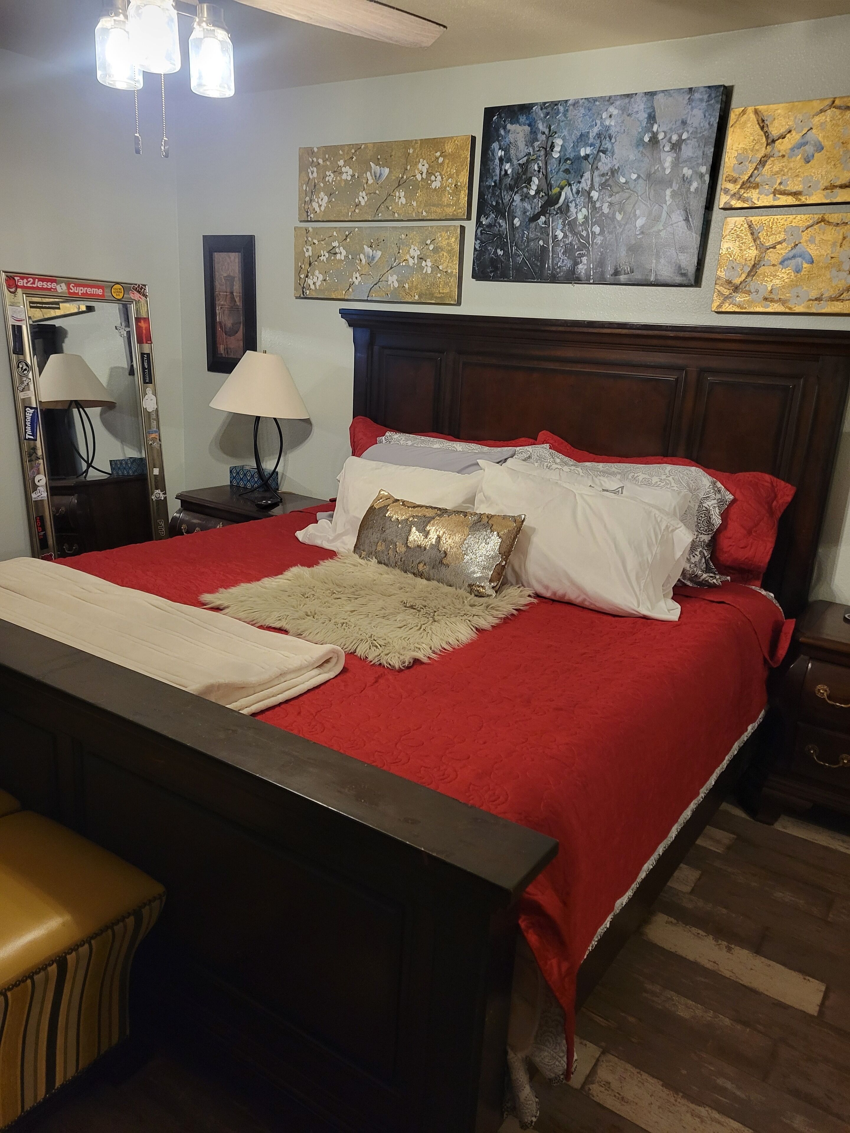 Una camera da letto con un letto in legno, biancheria rossa e cuscini decorativi. Si nota uno specchio con adesivi, una lampada e un'opera d'arte appesa al muro.