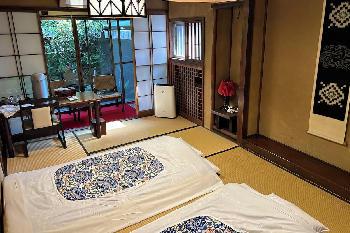 A traditional Japanese room with tatami flooring, a low table with a chair, a bed with patterned bedding, and sliding doors leading to a garden.