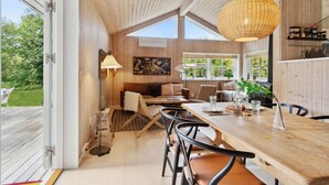 A cozy interior with a wooden ceiling, a dining area with a table and chairs, a sofa, and a large window providing natural light.