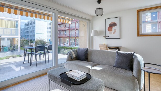 A modern living room with a grey sofa, a small round table, and a view of a residential building through large windows. - Nice Wallisellen 2br w/ Storage & W/D, nr Local Market, by Blueground (Zurich)