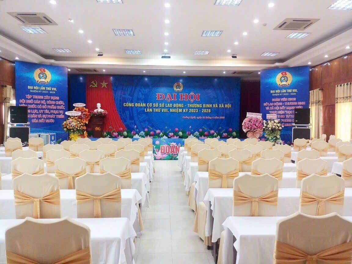 Un salón de banquetes con filas de mesas y sillas, un escenario con un banner y arreglos florales.
