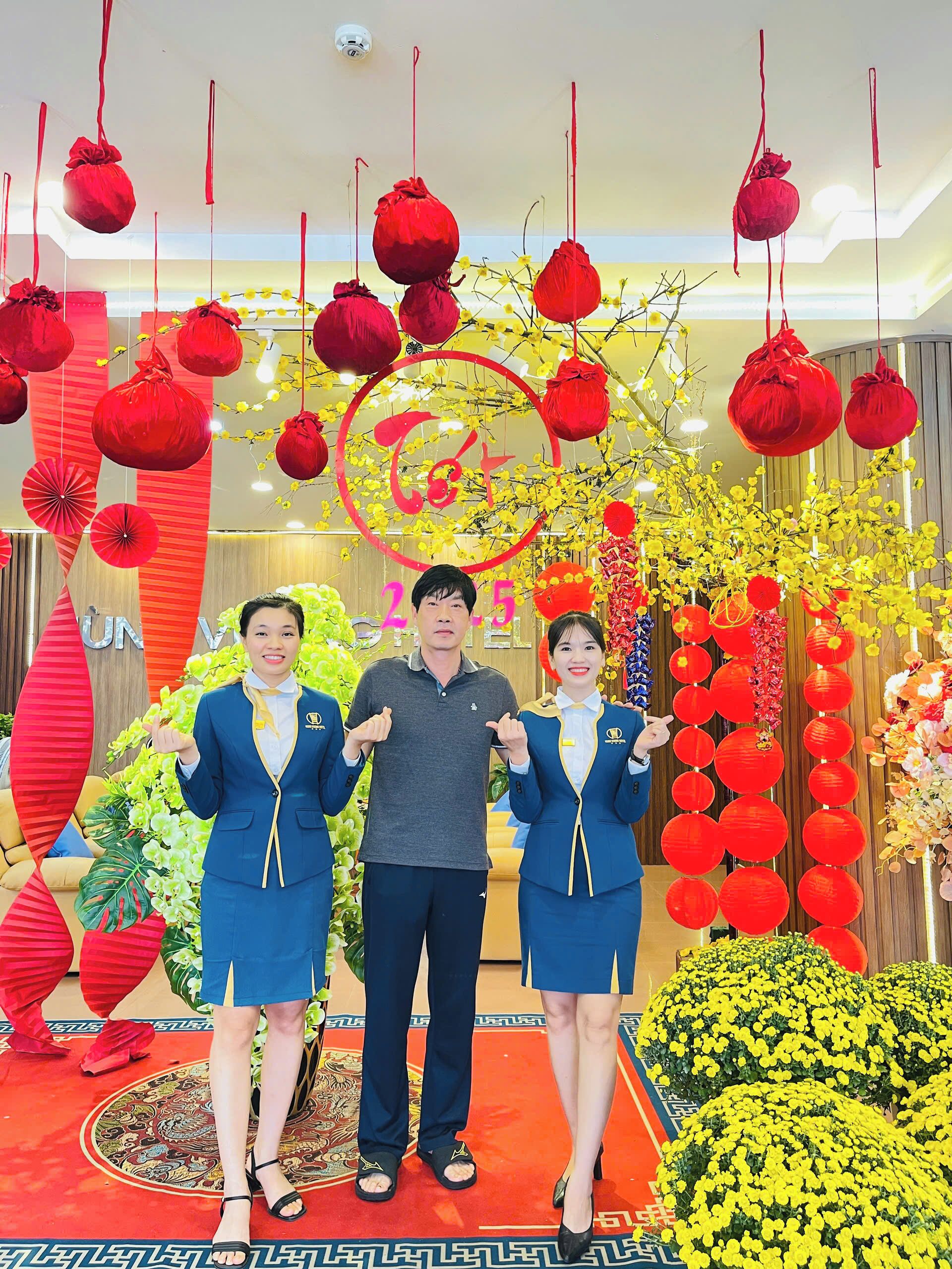 Three people standing in front of a festive decoration with hanging red lanterns and yellow flowers.