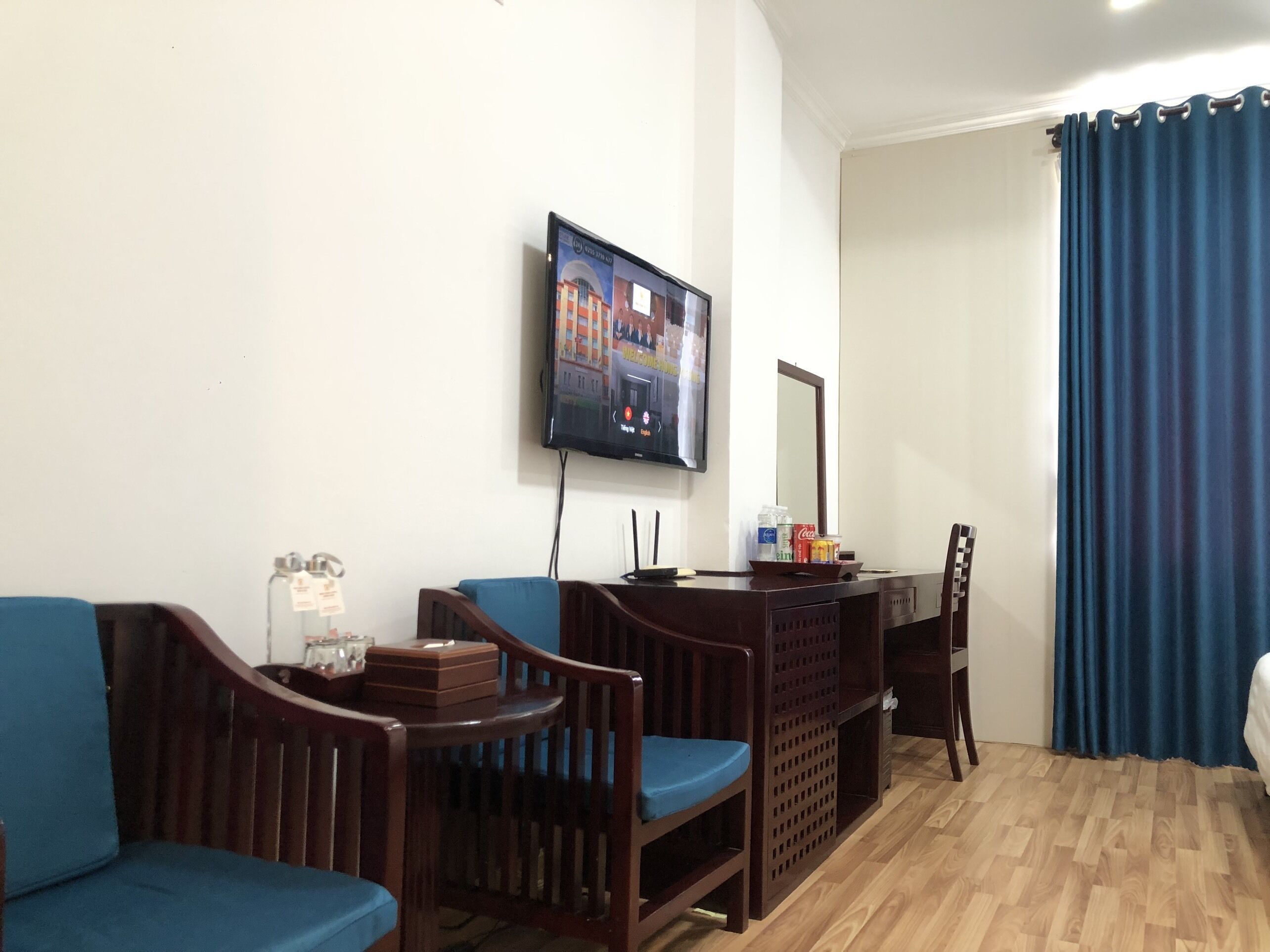 Ein Hotelzimmer mit einem Holztisch, zwei blauen Stühlen, einem an der Wand befestigten Fernseher und einem Bett mit blauem Vorhang.