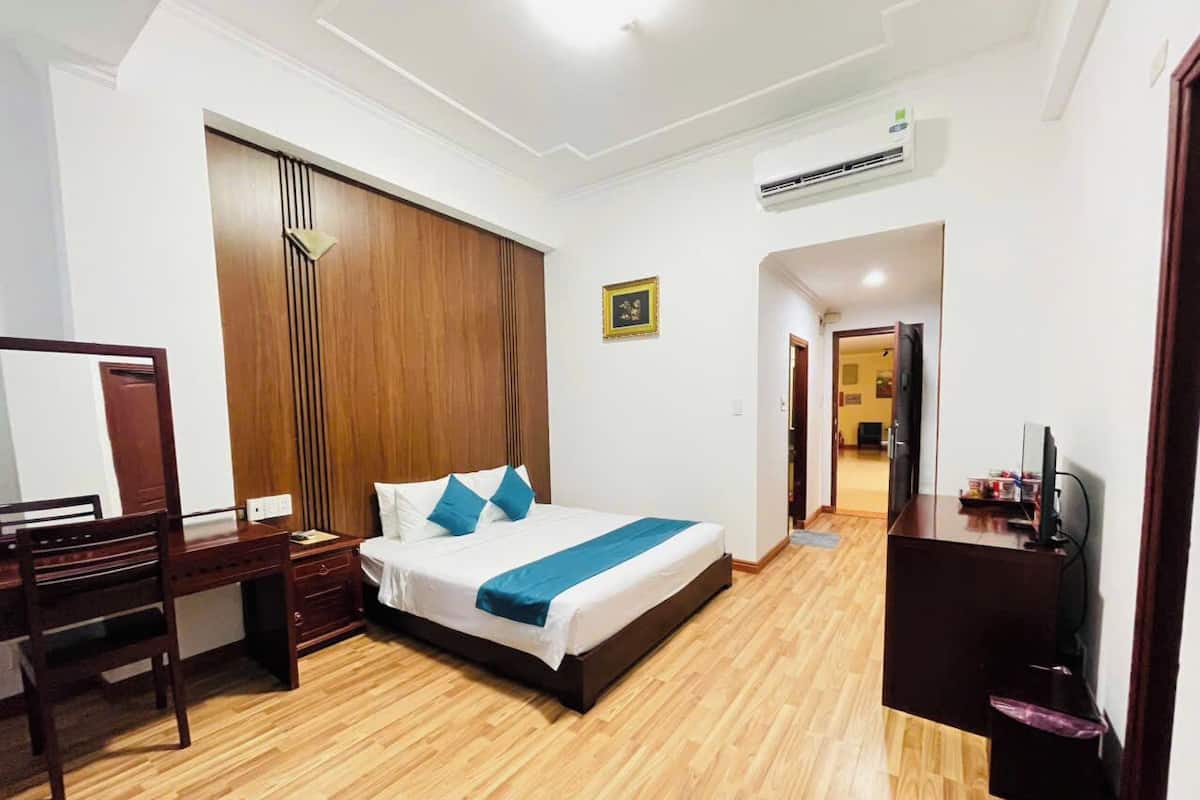 Habitación de hotel con suelo de madera, cama con colcha azul, escritorio con silla, armario y televisor sobre un mueble.