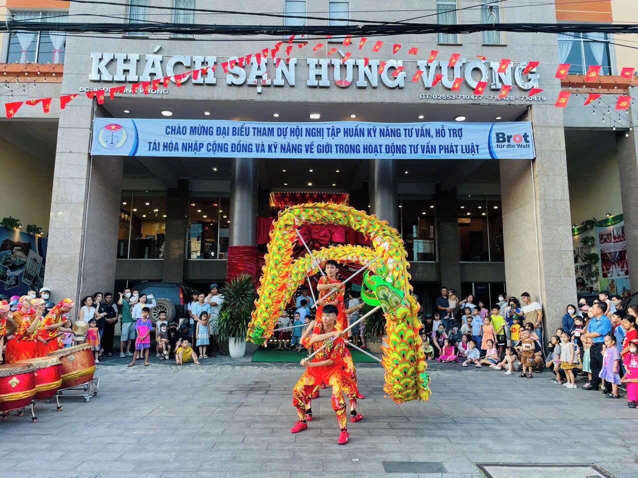 Una tradizionale esibizione di danza del drago di fronte a un edificio con un cartello che riporta la scritta "KHÃCHSÃN HUNG VUONG".