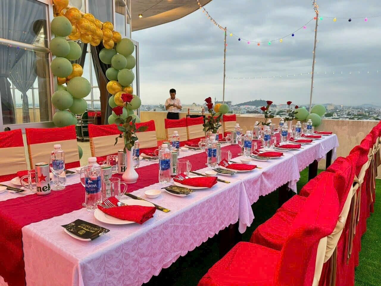 A rooftop dining area with tables set for a meal, decorated with balloons and flowers, and overlooking a cityscape.