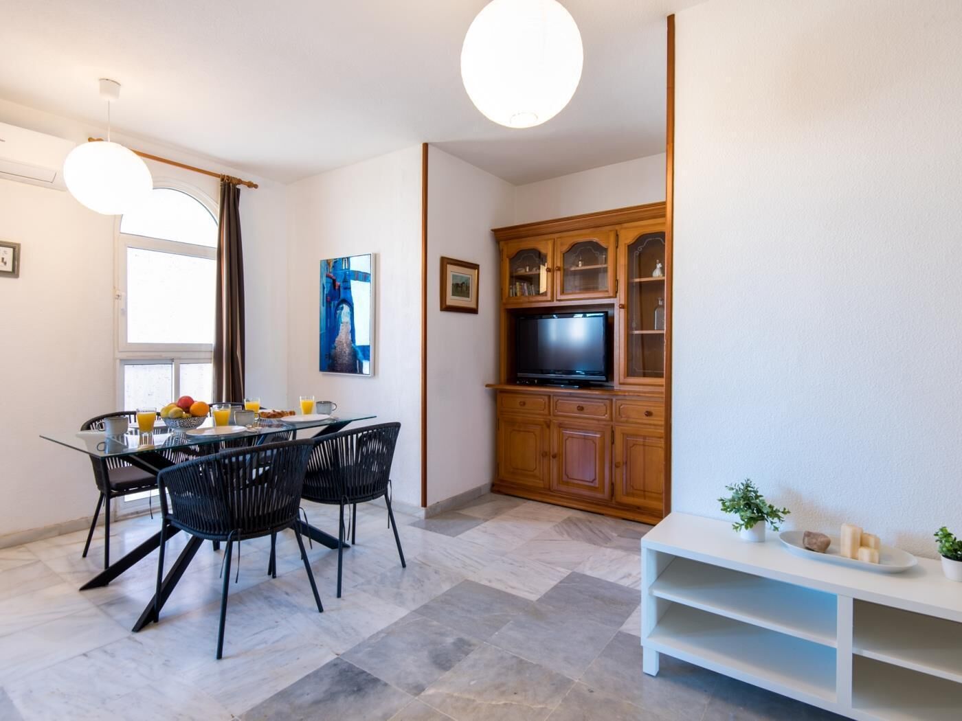 A modern dining area with a glass table and black chairs, a wooden cabinet with a television, and a white shelf with decorative items.