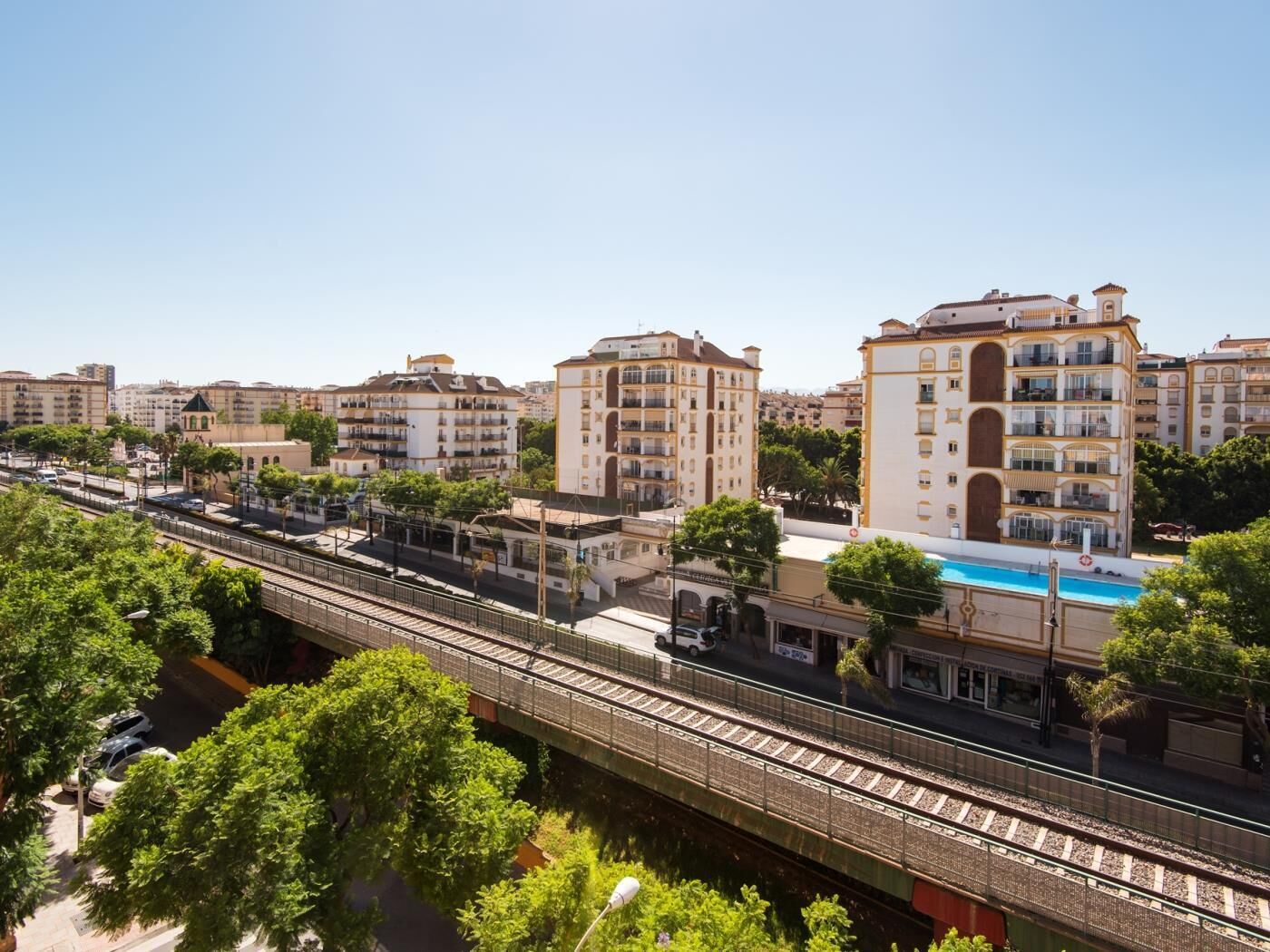 Un quartier urbain moderne avec un pont piétonnier enjambant une voie ferrée, entouré d’immeubles résidentiels à plusieurs étages et d’une piscine.