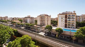 Un quartier urbain moderne avec un pont piétonnier enjambant une voie ferrée, entouré d’immeubles résidentiels à plusieurs étages et d’une piscine.