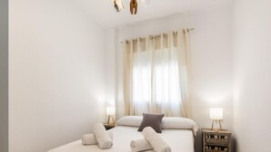 Una cama bien tendida con toallas enrolladas, una lámpara de noche y una pequeña mesita de madera con lámpara.