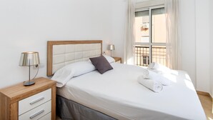 Una cama bien hecha con sábanas blancas y un almohadón oscuro, una mesita de noche con una lámpara y una ventana con cortinas.