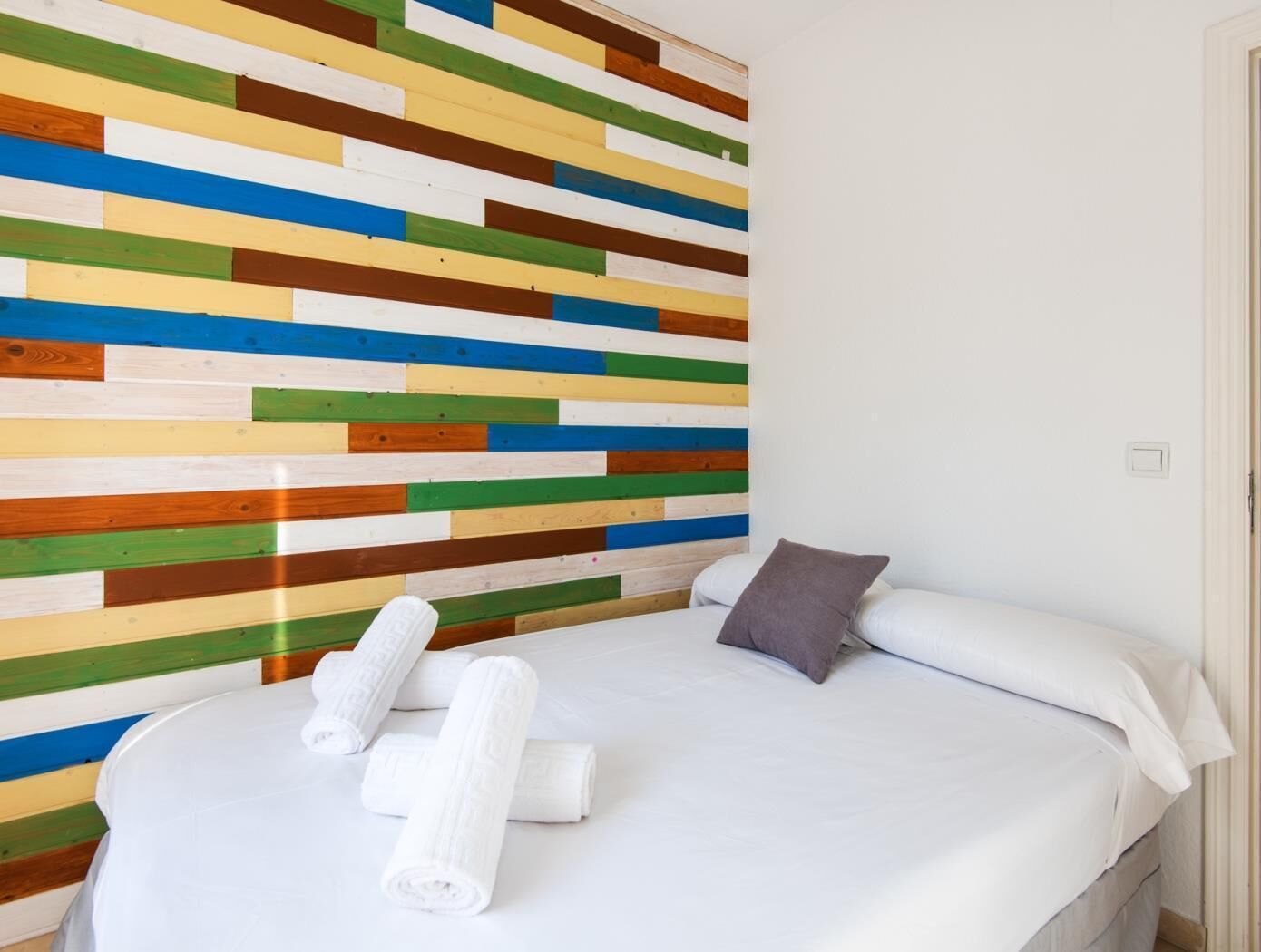 Una habitación de hotel con cama, ropa de cama blanca, almohada gris y pared a rayas coloridas.