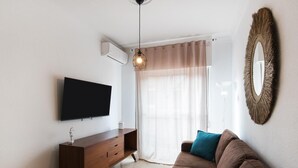 Ein modernes Wohnzimmer mit Flachbildfernseher, einem Holz-TV-Möbel, einem braunen Sofa mit türkisfarbenen Kissen, einer Pendelleuchte und einem großen Spiegel an der Wand.