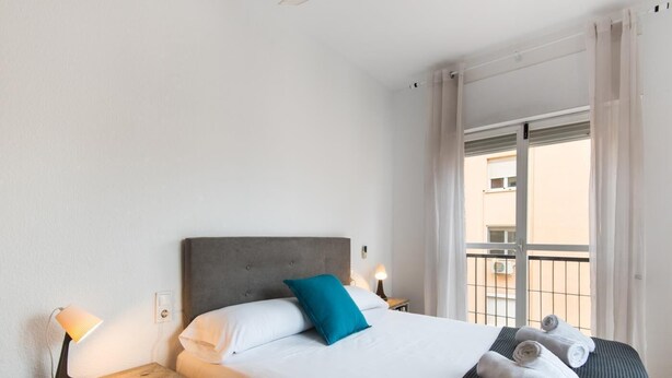 Ein ordentlich gemachtes Bett mit grauem Kopfteil, weißer Bettwäsche und einem blauen Kopfkissen. Zwei Nachttischlampen, ein Nachttisch mit Körben und ein Fenster mit Blick auf einen Balkon.