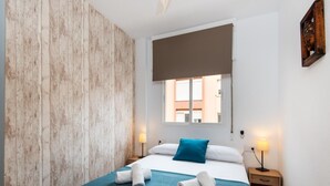 Ein modernes Hotelzimmer mit einer Wand in Holzoptik, einem großen Bett mit türkisfarbener Bettwäsche, zwei Nachttischlampen und Blick auf Gebäude durch ein Fenster.