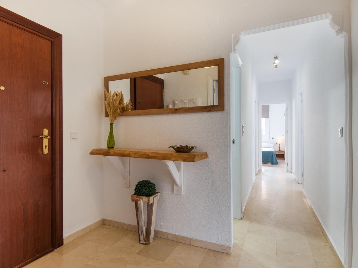 Un couloir avec une porte en bois, un miroir, une petite plante et une étagère sur laquelle se trouve un bol.