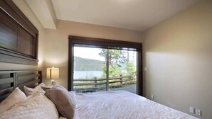 A bedroom with a large bed, a nightstand with a lamp, and a view of a lake through a glass door.