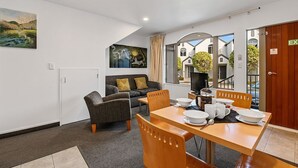 Een moderne kamer met een eettafel voor vier personen, een bank met kussens, een schilderij aan de muur en uitzicht op buiten door grote ramen.