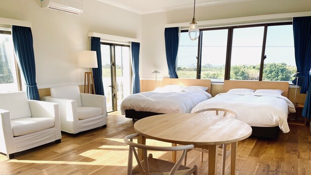 A hotel room with two beds, a round wooden table, two white armchairs, and a large window with curtains. - AZZURRA (Miyakojima)
