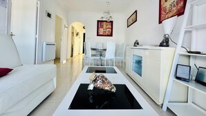 Ein modernes Wohnzimmer mit einem weißen Sofa, einem Couchtisch mit Dekoration, einem Essbereich mit weißen Stühlen und einer Küchenzeile mit weißen Schränken sowie einem Fernseher.