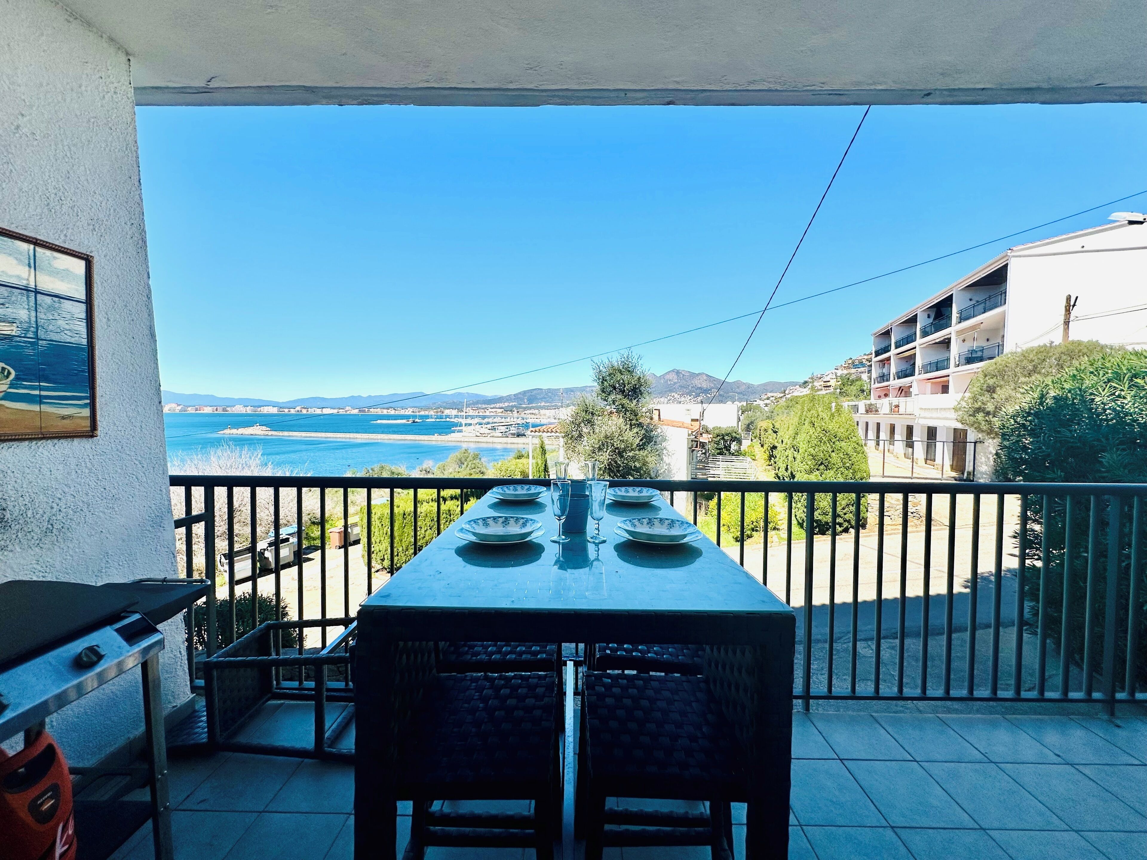 A balcony with a table set for dining, overlooking a coastal view with buildings and greenery.