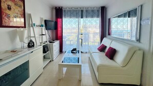 Ein Wohnzimmer mit weißem Sofa, Couchtisch, Fernseher auf einem Schrank, einer Leiter und einem Fenster mit Blick.