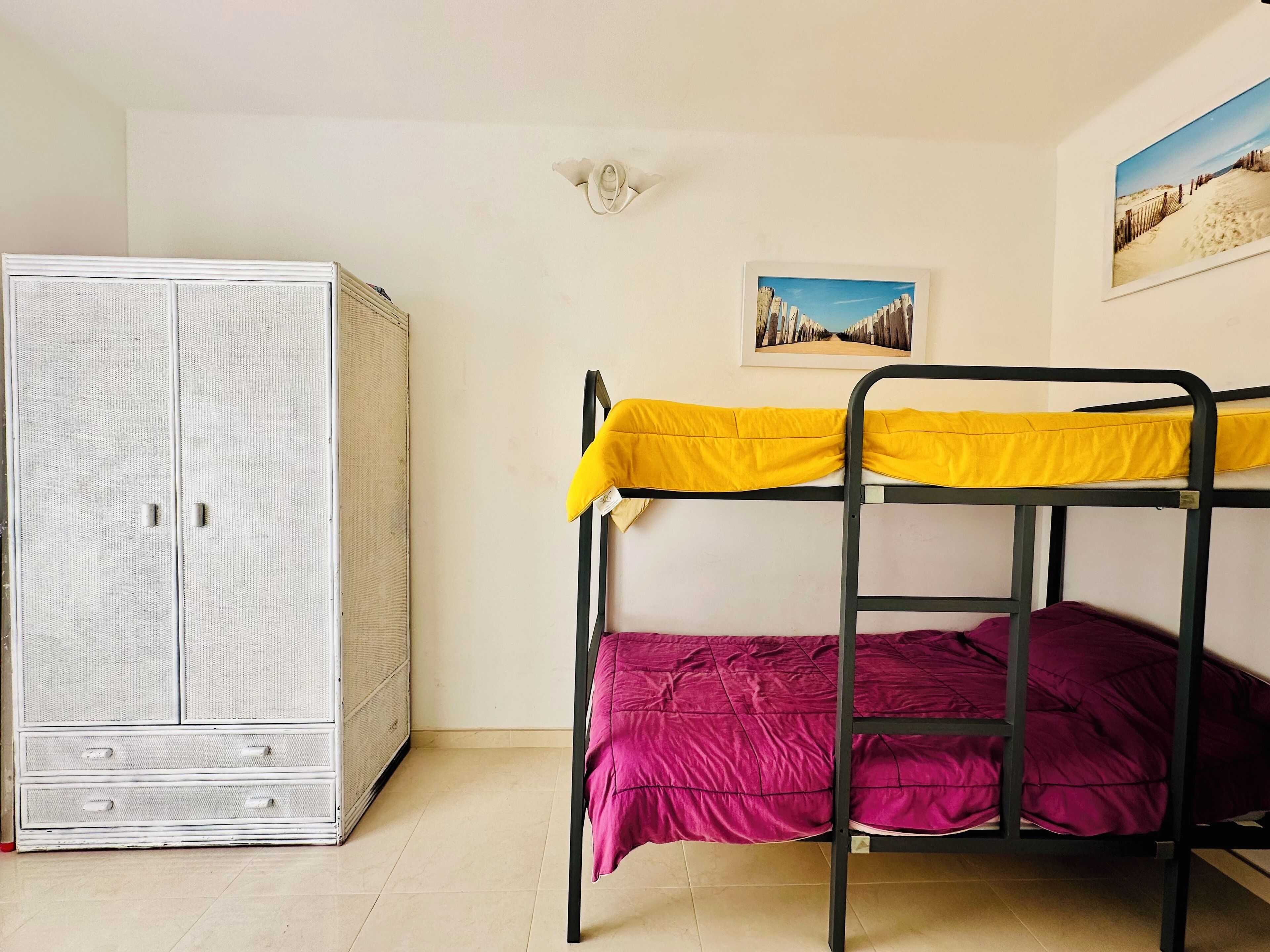 A bunk bed with yellow and pink bedding, a wardrobe with two drawers, and a framed picture on the wall.