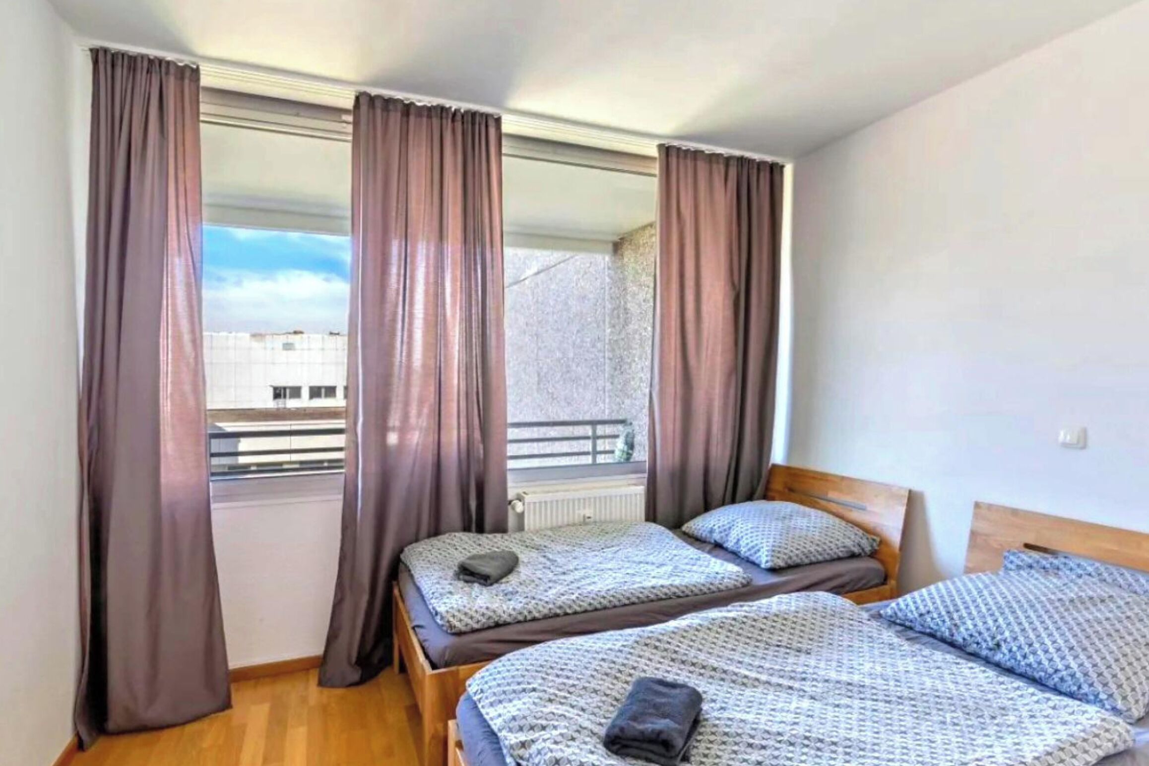 Ein Zimmer mit zwei Betten, einem fenster mit Vorhängen und Blick auf Gebäude.