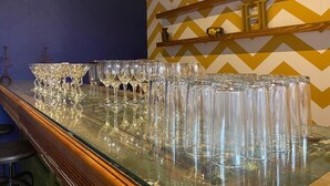 Una zona de bar con una barra alargada que exhibe numerosos vasos vacíos, un estante de madera con objetos decorativos y una pared en el fondo con estampado en zigzag.