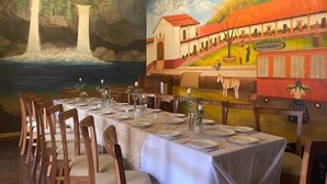 Un comedor con mesas dispuestas para una comida, un mural en la pared que representa una escena campestre y una pintura de cascada sobre las mesas.