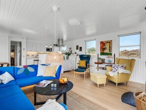 Una sala moderna con un sofá azul, un sillón amarillo y un comedor con muebles blancos.