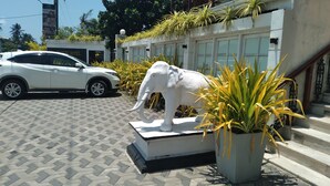 Un coche blanco estacionado frente a un edificio con una gran estatua de elefante y plantas en macetas.