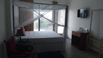 A single bed with a canopy, a desk with a chair, a television on the wall, and a view of palm trees outside.