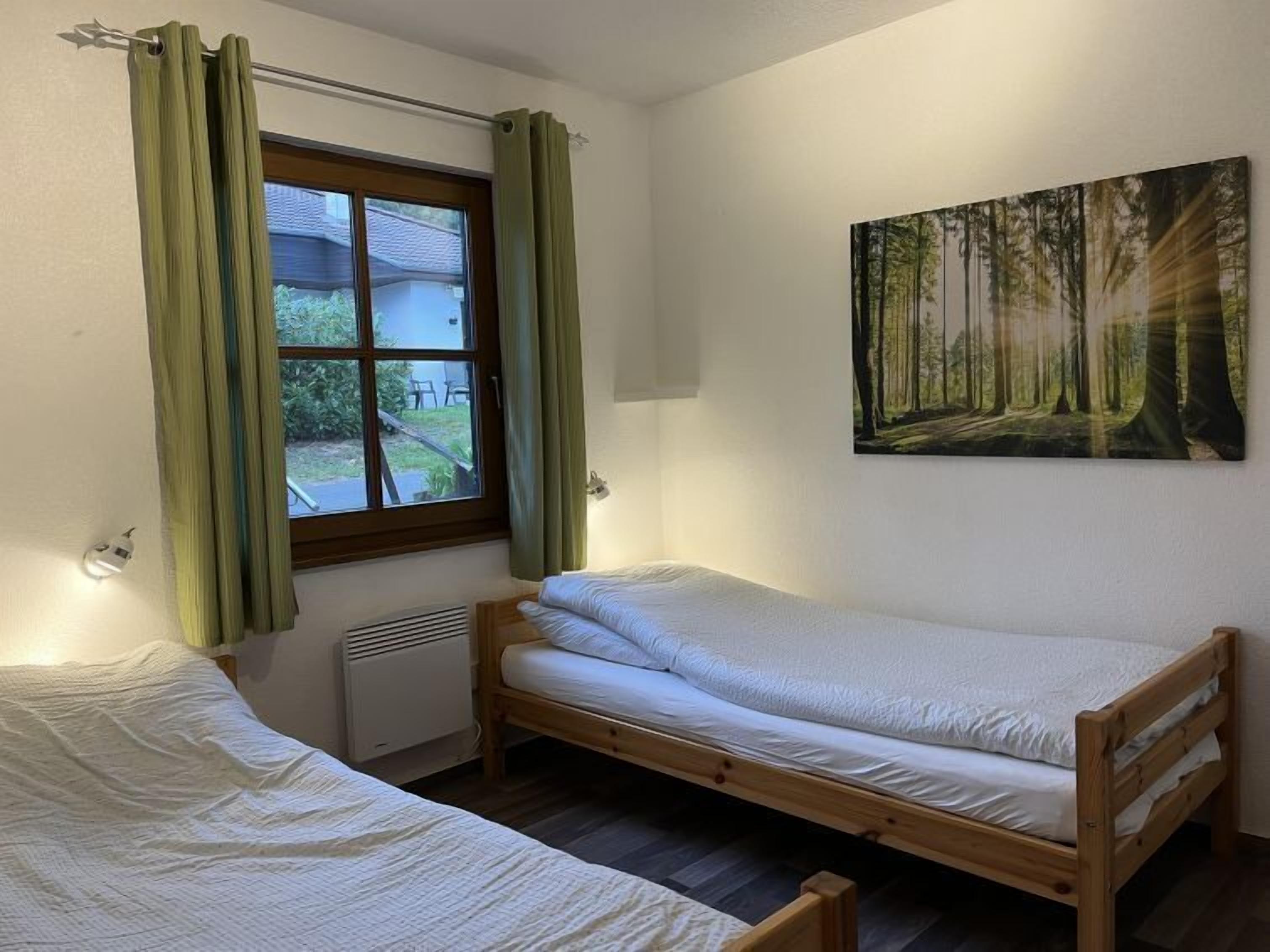 Un petit logement composé d’une seule pièce, avec un lit en bois, une fenêtre avec des rideaux, un radiateur et un tableau sur le thème de la forêt accroché au mur.