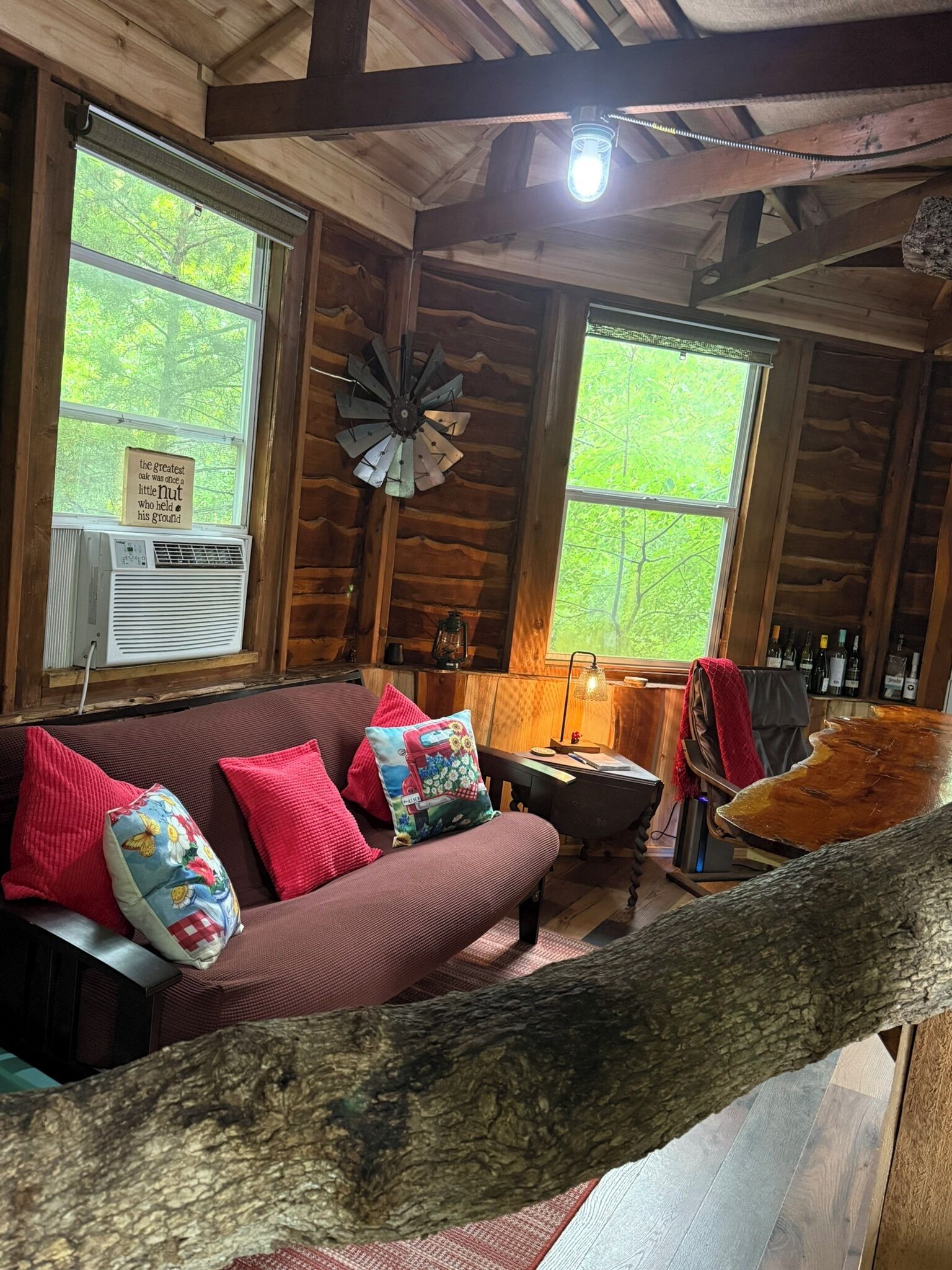 A cozy cabin interior with a wooden ceiling, a sofa with colorful cushions, a clock, and a window with a view of greenery.