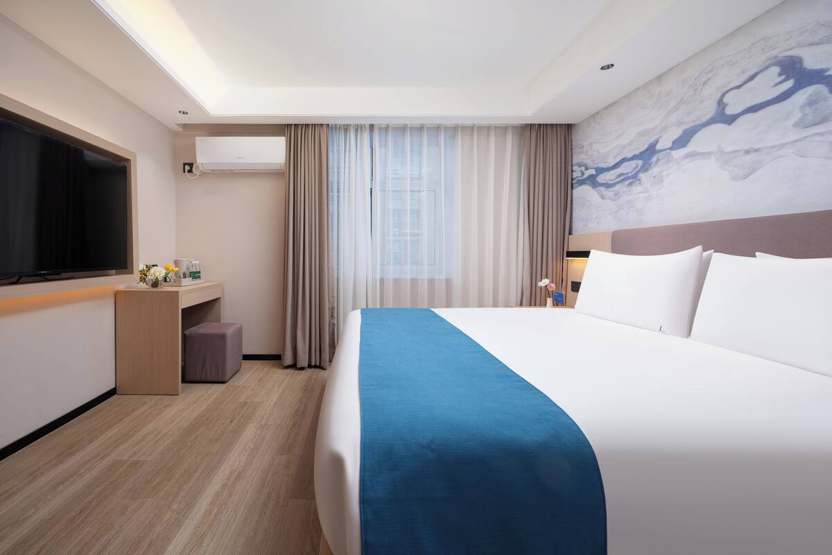A hotel room with a large bed, a desk with a flat-screen TV, a window with sheer curtains, and a marble wall.
