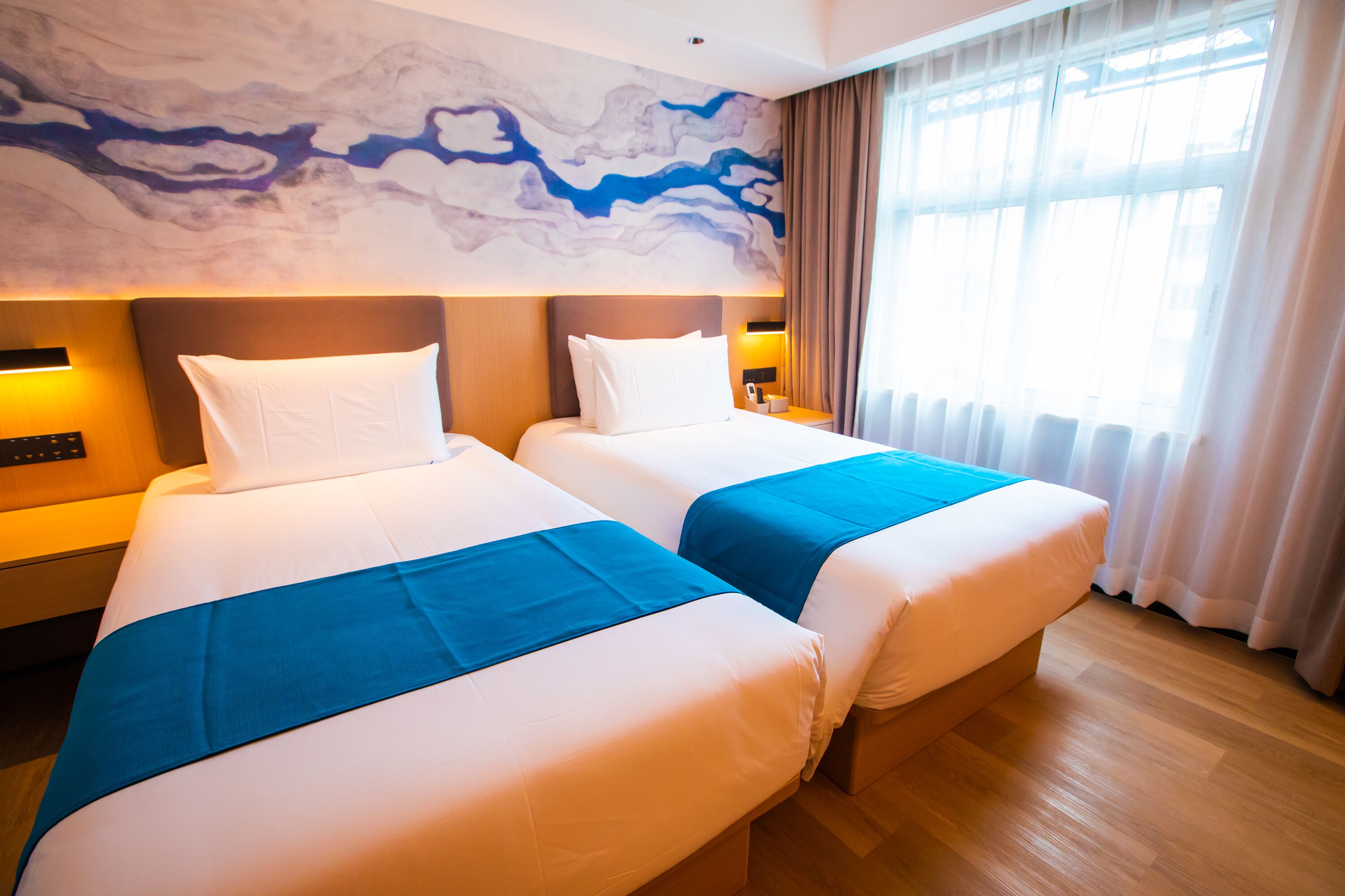 Habitación de hotel con dos camas, un mural de mármol, un ventanal con cortinas y suelo de madera.