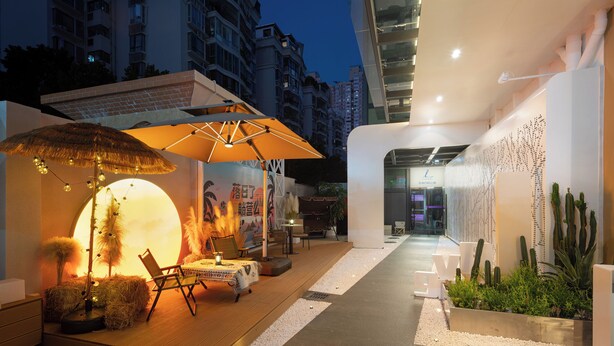 An outdoor seating area with a thatched umbrella, a round table, and a chair. There is a wall with a cactus garden and a building entrance in the background. - NAIVE L Executive Apartments Coast City (Shenzhen)