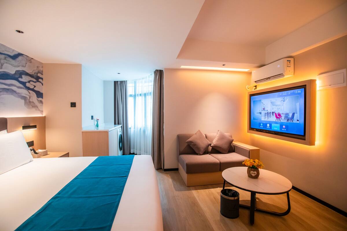 A modern hotel room with a bed, a sofa, a small table with a vase, a TV on the wall, and a window with curtains.