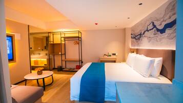 A hotel room with a bed, a desk, a chair, a small table with a vase, a bathroom with a mirror, and a wall with a painting.