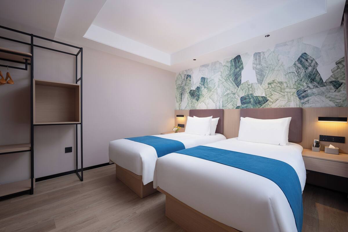 A hotel room with two beds, a wooden floor, a wall with a textured mural, and a skylight.